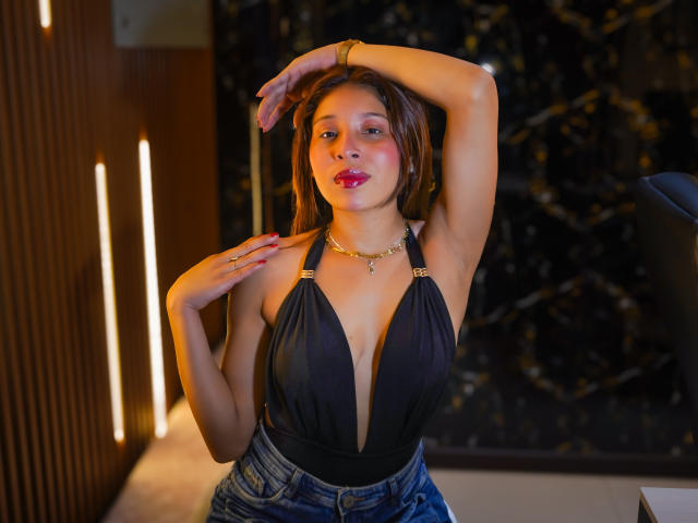 JaneKetty with medium tits, wearing a deep black halter top and blue jeans, has her arm raised, brown hair, red lipstick, gold necklace, and a ring on her right hand.