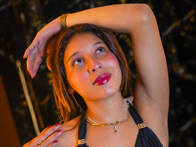 JaneKetty with shiny red lipstick, arm raised, black halter top, gold necklace, red nails, brown hair, looking up, glowing orange light, black background with gold sparkles.