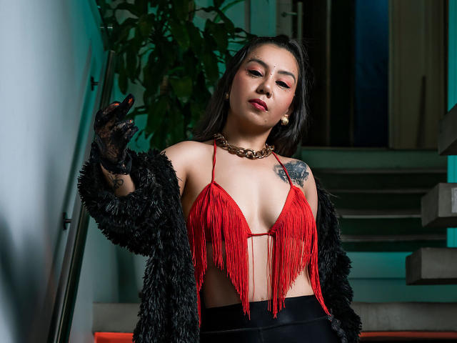 MissCharlotteJolie in a red fringe top, black furry jacket, black gloves, gold chain, and black pants, with a tattoo on her left shoulder, looking confident.