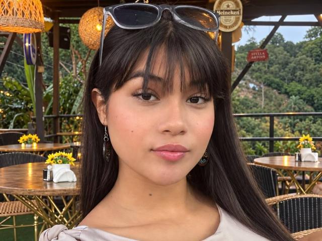 AshlyLee with long black hair, bangs, and sunglasses on her head, wearing a white top, has a small mole on her left cheek, and a subtle smile.