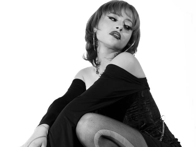 ViolettClay with short, wavy hair, off-shoulder black dress, and black tights, sitting, looking down with half-closed eyes, wearing hoop earrings and a necklace, in black-and-white.