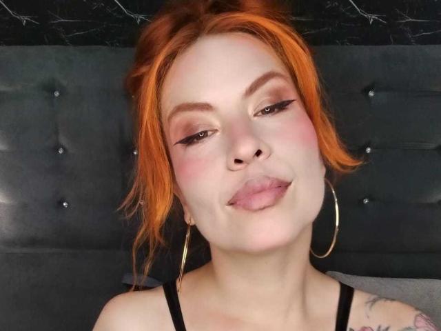LilithSnop met oranje haar, roze wangen en gouden oorringen, die haar tong uitsteekt met een lichtelijk zelfvoldane uitdrukking, draagt een zwarte top en heeft een kleine tatoeage..