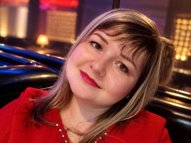 KellyMary tiene el cabello rubio, lápiz labial rojo y una sonrisa encantadora, lleva una camisa roja con tachuelas y está ligeramente inclinada hacia un lado, dentro de un coche por la noche..