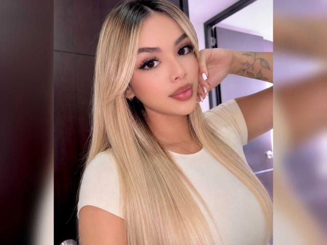 ZoeVibe has long blonde hair, full lips, and a tattoo on her arm. She's wearing a white shirt, looking at the camera with a relaxed, sexy expression.