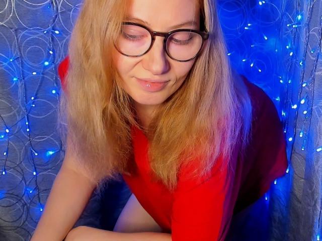 PaulinaSunny with blonde hair, glasses, and red top, crouching, smiling, and showing a small, shiny lip piercing. Blue lights in background.