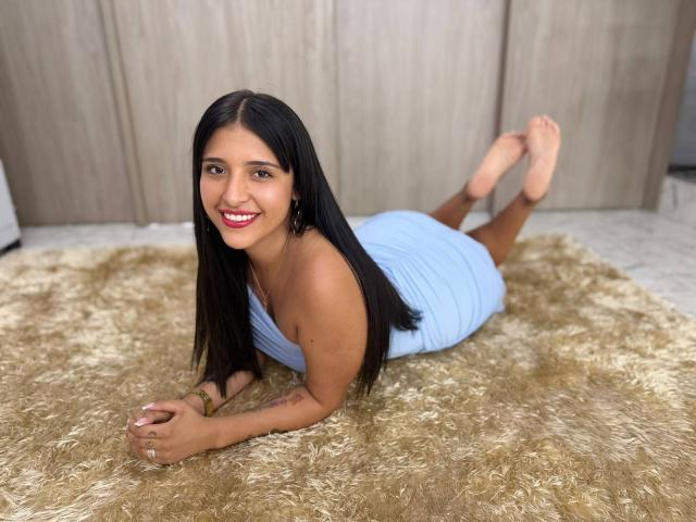 SophieCan lying on a shaggy beige rug, wearing a light blue dress, smiling with red lipstick, black hair, and hoop earrings, showing her bare ass and feet in the air.