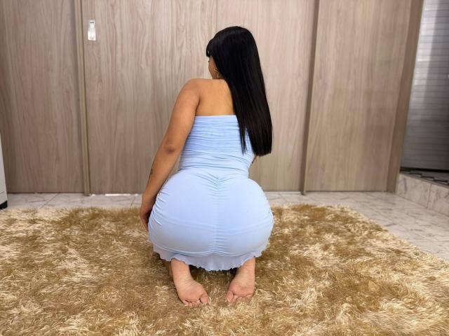 SophieCan is kneeling on a fluffy beige rug, wearing a tight, light blue dress that highlights her big ass and bare feet. She has long black hair.