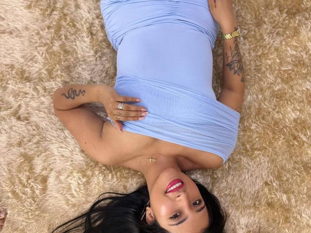 Sophie, deitada em um tapete fofo, vestindo um vestido azul justo que revela seios pequenos, com cabelo preto, batom vermelho, tatuagens e um relógio de ouro, sorrindo para a câmera..