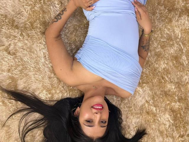 SophieCan is lying on a fluffy beige rug, wearing a tight blue dress that reveals her tits, with long black hair, red lipstick, and tattoos on her arms.