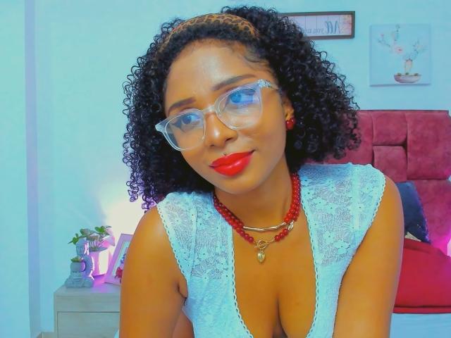 MeganWlliams with curly black hair, wearing clear glasses, red lipstick, white lace top, red bead necklace, and red earrings, showing her cleavage. Bedroom setting.