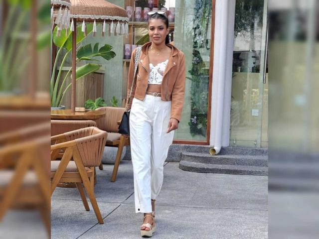 CamilaWest, usando uma jaqueta marrom, calças brancas de cintura alta, sandálias marrons e uma blusa branca com estampas florais, caminhando ao ar livre com uma bolsa preta, cabelo castanho preso em um rabo de cavalo, sorrindo..