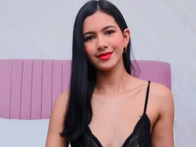 AmilyPreston has long black hair, red lipstick, and wears a black lace top. She's smiling with red blush on her cheeks. Her small tits are visible.