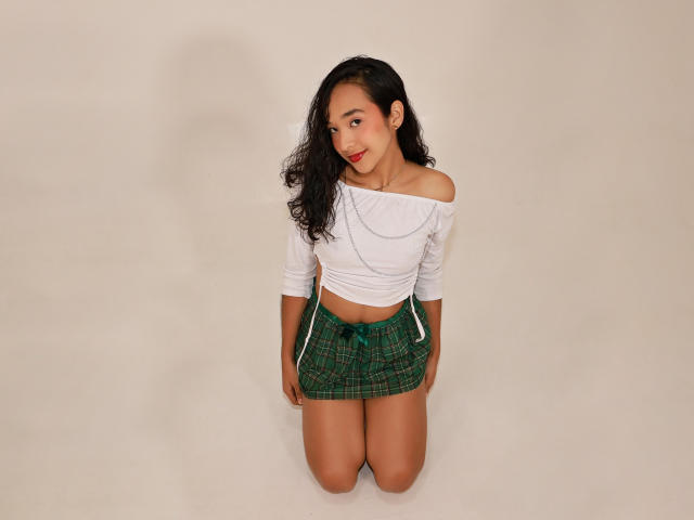 Whitneypacee kneeling, wearing a white off-shoulder top and green plaid mini-skirt, with long curly black hair, red lipstick, and a cute smile, on a beige background.