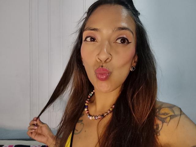 IsaChaud, con le labbra corrugate e un anello al naso, tiene i capelli castani, indossa una collana colorata di perline, ha tatuaggi sulle spalle, occhi marroni, un trucco leggero, indossa una maglietta gialla, orecchini, illuminazione soffusa, sorride..