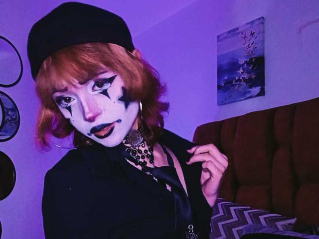 ViolettClay con i capelli rossi, il viso dipinto di bianco, l'eyeliner nero e il rossetto blu, indossa un berretto nero, una giacca e una collana a stampa leopardo, tenendo la sua camicia. Stanza di colore viola..