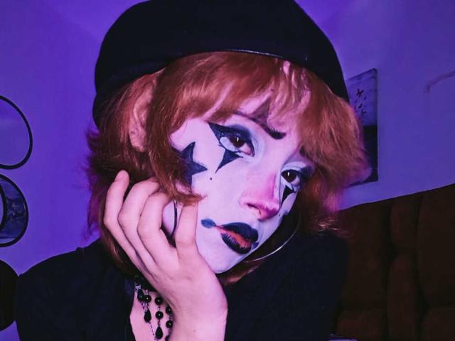 ViolettClay heeft oranje haar, witte gezichtsbeschildering met zwarte sterren en vleugelmake-up, zwarte lippenstift en een zwarte muts. Ze raakt haar wang aan met een zwarte armband met kralen..
