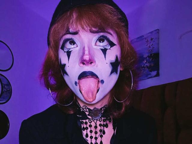 ViolettClay avec du maquillage blanc sur le visage, des étoiles noires, la langue tirée, portant une casquette noire, des boucles d'oreilles cerceaux et un collier noir, sous un éclairage violet. Cheveux bouclés roux..