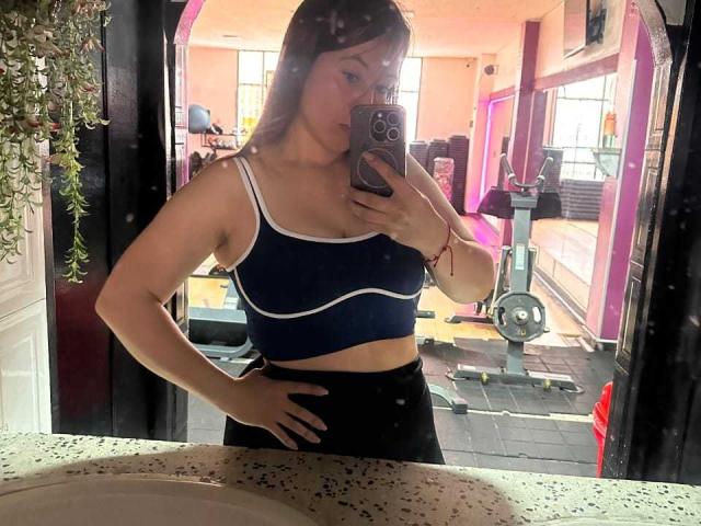 LizBellaa with medium tits in a blue sports bra and black high-waisted shorts, taking a mirror selfie in a gym, holding a pink phone, hand on hip.