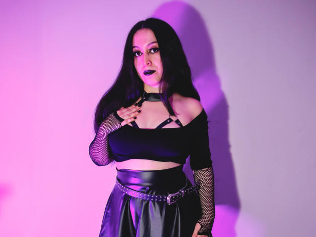 KendraSimons with dark lipstick, black mesh sleeves, strappy top, and shiny black skirt, hand on chest, purple and pink lighting, long black hair, confident expression.