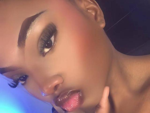 Mulher negra atraente com olhos castanhos brilhantes, sobrancelhas grossas, blush rosa e lábios rosados levemente entreabertos, tocando o queixo com o dedo, olhando sedutivamente para a câmera..