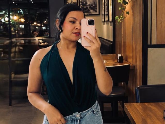 MelanyPorto with dark green, deep V-neck top showing her ass, holding a pink phone, wearing blue jeans, taking a mirror selfie in a dimly lit cafe.