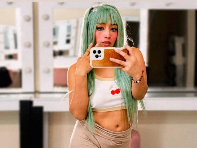 AilenRosse, com longos cabelos verdes, vestindo uma cropped branca com uma cereja, tirando uma selfie no espelho com um celular, mostrando seus pequenos seios e abdômen tonificado..