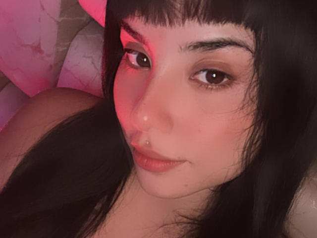 GraceClarke with dark hair, brown eyes, and a nose piercing, looking at the camera with a slight smile, pink light on the left side of her face.