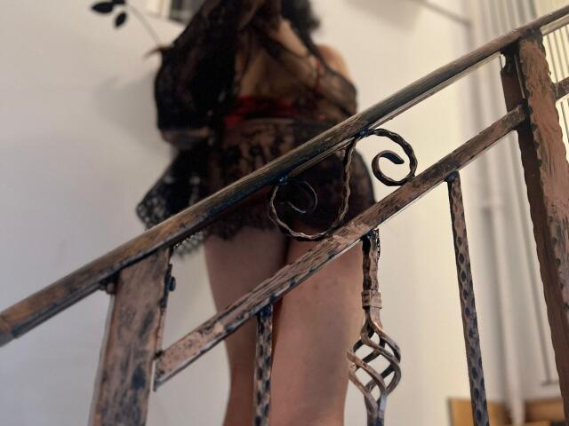 IrisIrs in sheer black lace lingerie with visible ass and legs, standing on a rusty metal staircase, blurred head, curly dark hair, white wall in background.