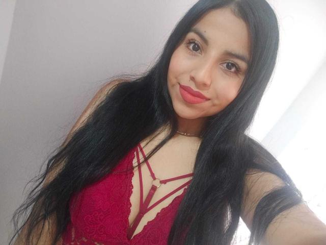 EmmaSollins with long black hair, red lace bra, and strappy design, showing her tits, smiling with red lipstick, wearing a gold necklace, in a bright room.