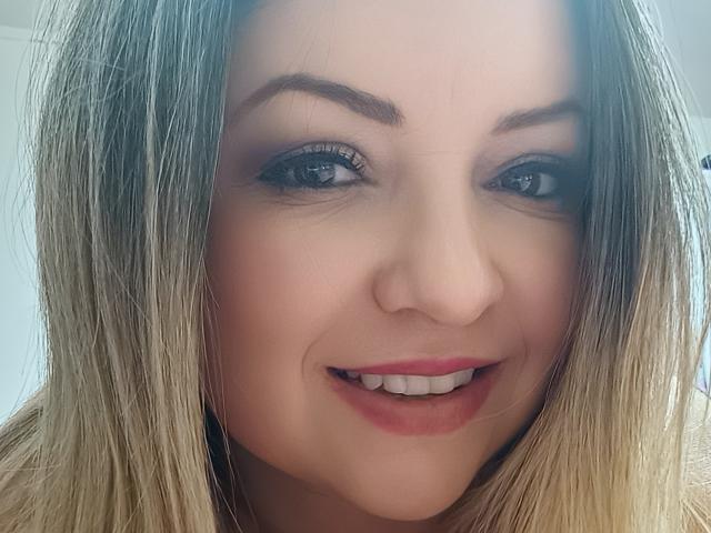 NatalyHami's face, showing blonde hair, dark eyebrows, blue eyes, and a slight smile with red lipstick, has a cute and sexy look, featuring her ass in the background.