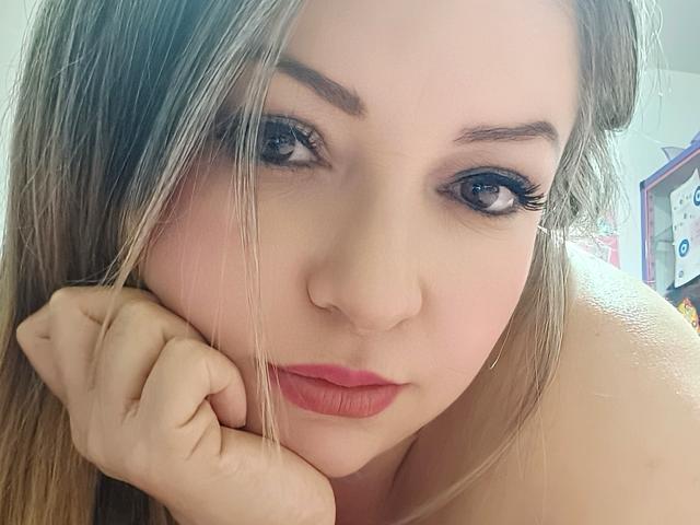 NatalyHami has blonde hair, blue eyes, and is looking at the camera with her hand under her chin, wearing pink lipstick. Her tits are not visible.