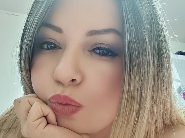 NatalyHami has pouty pink lips, dark blue eyes with black eyeliner, and blonde hair. She's resting her chin on her hand, looking at the camera.