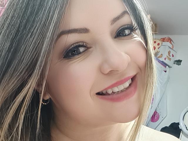 NatalyHami's face, smiling with pink lipstick, blue eyes, and brown hair. She's got a cute smile, small gold earrings, and her ass isn't visible in this shot.