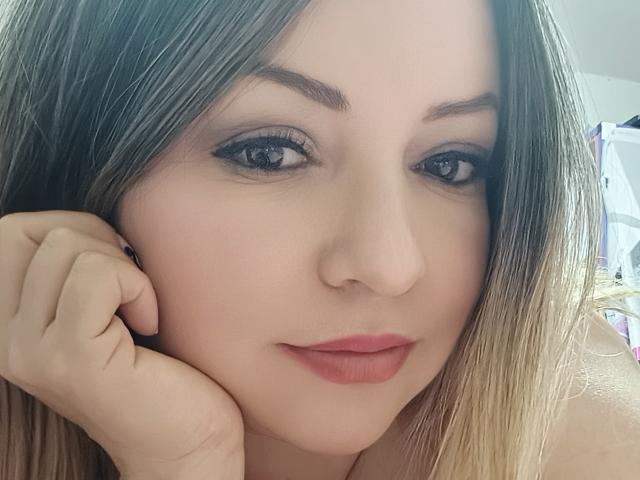 NatalyHami has light skin, brown eyes, and blonde hair, with her hand on her cheek, showing pink lips and subtle makeup. Her tits are not visible.