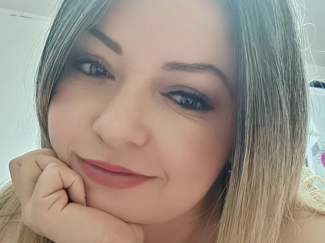 NatalyHami has blonde hair, blue eyes, and is smiling with pink lips. She's resting her chin on her hand, looking cute and sexy. Her makeup includes dark eyeliner.