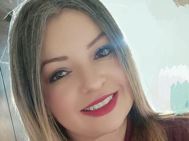 NatalyHami's face with light brown hair, blue eyes, and red lipstick, smiling with straight white teeth, looking at the camera, soft sunlight on the left side of her face.