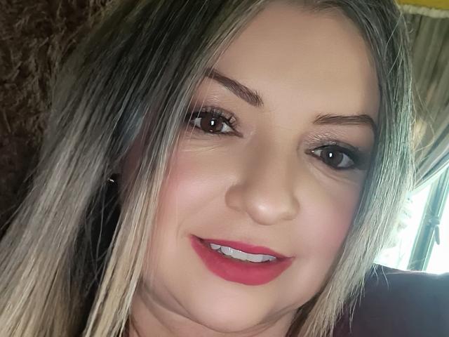 NatalyHami's face, with blonde hair, brown eyes, red lipstick, and slight smile, showing her white teeth. She has light skin and is looking at the camera.