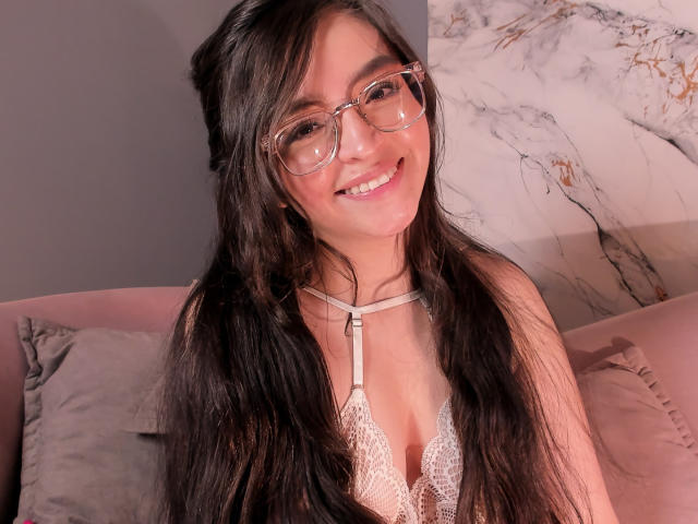 Eva McAllister mit langem, schwarzem Haar, Brille, lächelnd, trägt einen Spitzen-BH, zeigt ihre kleinen Brüste, sitzt auf einem rosa Sofa, eine Marmorwand ist im Hintergrund zu sehen..