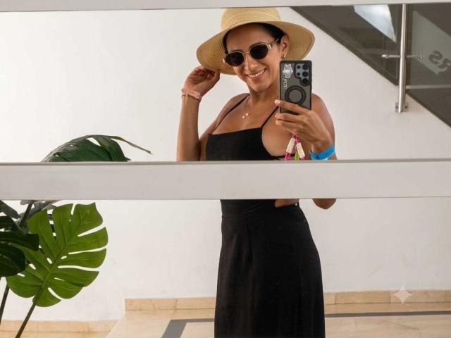 JadeFoster, à la peau foncée, portant une robe noire, un chapeau de paille et des lunettes de soleil, tenant un téléphone, souriante, se montrant les seins dans un selfie devant un miroir. Une plante est visible en arrière-plan..