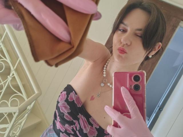 BerryDiva con i capelli castano corti, che indossa guanti rosa, un abito floreale senza spalline, una collana di perle, che fa l'occhiolino, tiene in mano un telefono rosa, selfie allo specchio, una borsa marrone in primo piano, guanti rosa che tengono la borsa..