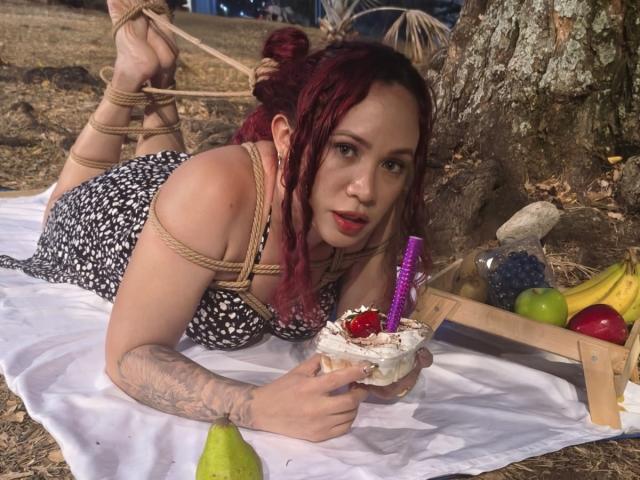 SophieSweetst with red braids, rope bondage, lying on white blanket, eating cake with purple candle, tattooed arm, black and white dress, surrounded by fruit and wooden stand.