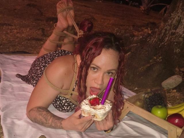 SophieSweetst, com cabelo ruivo, usando amarras de corda, comendo bolo com uma vela roxa, braço tatuado, deitada em um cobertor branco, com uma cesta de frutas ao lado dela..