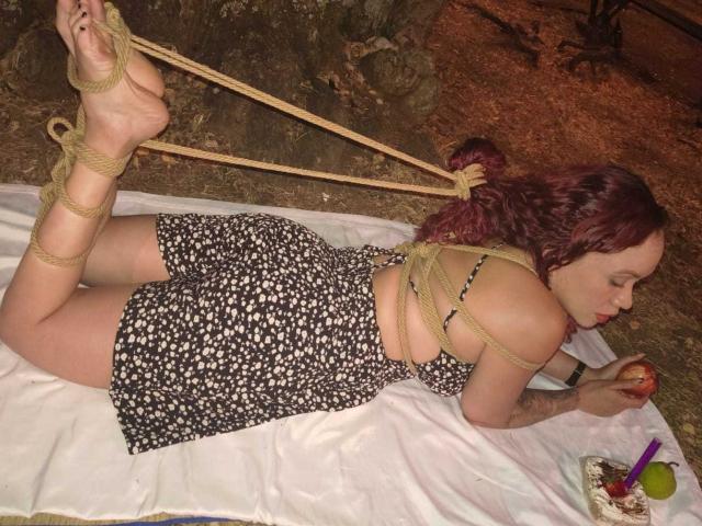 SophieSweetst liegt auf einer weißen Decke, mit Seilen gefesselt, trägt ein schwarzes Kleid mit Blumenmuster, hat rotes Haar und isst einen Apfel. Ihr Hintern und ihre Füße sind gefesselt..