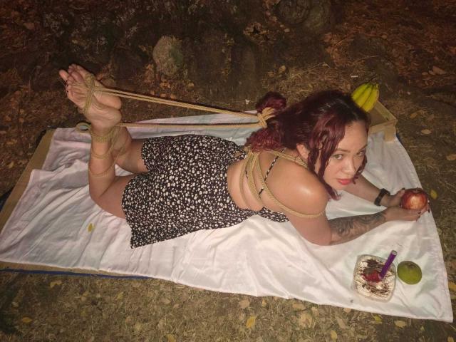 SophieSweetst está atada con una cuerda, acostada sobre una manta, vestida con un vestido floral negro, con las nalgas levantadas, sosteniendo una manzana, cerca de un pastel con una pajita morada..