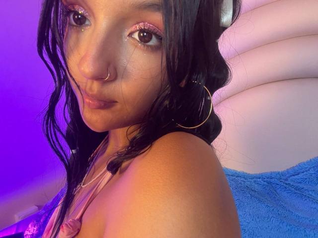 PaulineRey has dark wavy hair, brown eyes, pink glitter eyeshadow, nose ring, hoop earrings, and a subtle smile. She's in a pink top, with a blue towel.