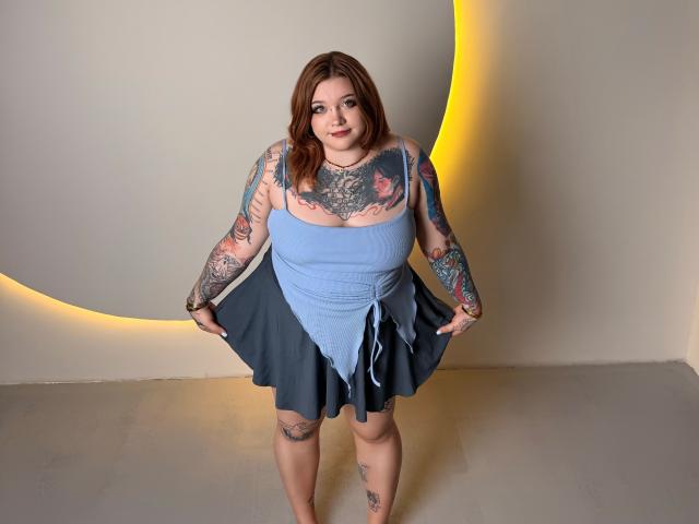 YoureGift, a plus-sized woman with tattoos, has brown hair, blue top, and gray skirt, smiling, holding skirt edges, showcasing tattoos on arms, chest, and legs.