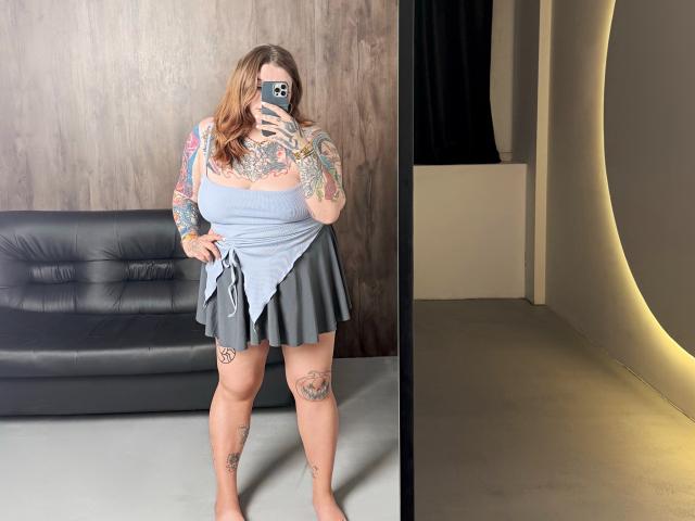YoureGift with tattoos, holding phone, wearing gray top and pleated skirt, standing barefoot, showing tattoos on thighs and arms, in front of black leather couch.