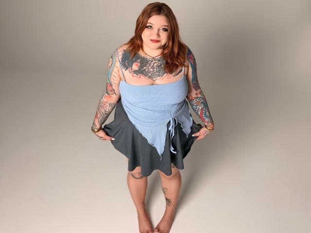 YoureGift, a plus-sized woman with tattoos, wearing a blue and gray dress, smiling, holding the skirt, standing barefoot, with red hair and light skin.