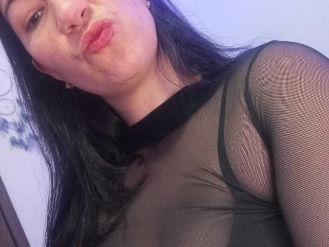 Yessi Soto, com os lábios curvados, blusa de malha preta que mostra os seios, cabelo preto longo, pele clara, brincos pequenos, fundo levemente azulado..