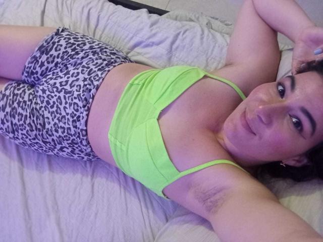 YessiSoto lying on a bed, wearing a neon green sports bra and leopard print shorts, armpits visible, smiling at the camera. Her tits are small, ass is cute.